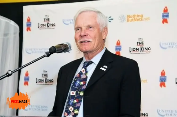 Ted Turner stands at a podium speaking into a microphone. The backdrop features logos for.