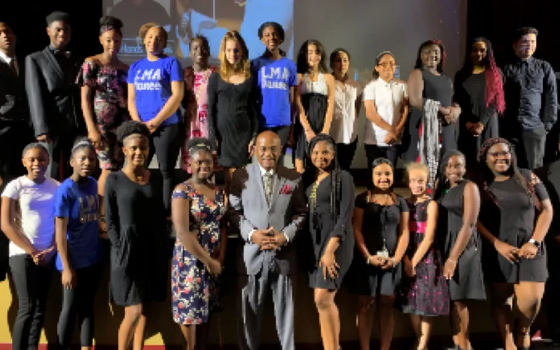 A group of young girls and adults posing on a stage, in a gray suit at the center  Willie Gary)  all smiling during a formal event.