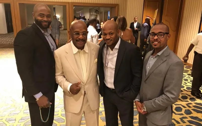 3 men in suits posing together at an indoor event, celebrity Willie Gary in a beige suit at the center smiling and standing.