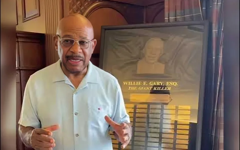 Willie Gary wearing glasses and a light shirt speaking indoors beside a plaque honoring Willie E. Gary,