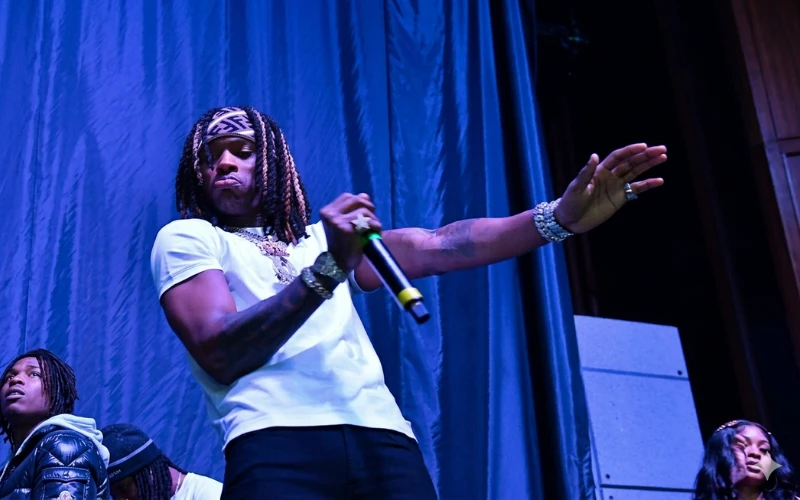 Rapper performing on stage with a microphone in hand against a dark blue curtain backdrop.