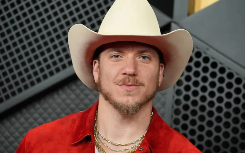 A close-up portrait of musician Brandon Lake wearing a light-colored cowboy hat, a red jacket, and gold chains