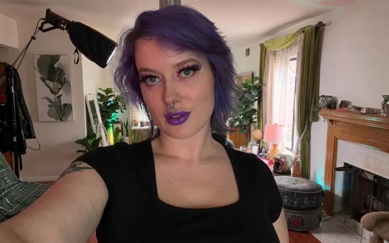 Nyx Baltimore with purple hair and bold makeup taking a selfie at home.